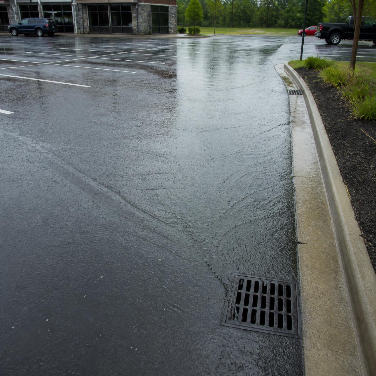 Rainwater sheeting across a properly sloped commercial asphalt parking lot toward a storm drain grate at the curb
